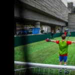 A group of people playing 5-a-side football on an outdoor field with artificial turf. The player in the foreground, dressed in a red jersey and green vest, stands with arms outstretched facing the goal. Urban buildings and a cityscape are visible in the background. © Powerleague