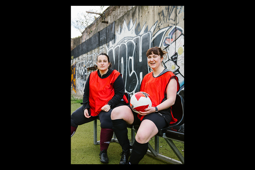 Two female soccer players in red jerseys sit on a bench against a graffiti-covered wall at the home of 5-a-side. One is holding a soccer ball, and both are dressed in black shorts and knee-high socks. They appear to be on an outdoor soccer field. © Powerleague