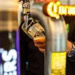 A person is pouring beer from a tap into a glass at a bar, their hands steady as they control the flow. The branding on the taps is partially visible. Wearing a dark long-sleeved shirt and a wristwatch, they work with precision. In the blurred background, there's chatter about function room hire. © Powerleague