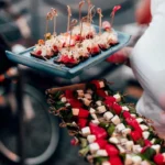 A person is holding two trays of assorted appetizers, perfect for any function room hire. The top tray features skewers with sprouts and red toppings, while the bottom tray showcases skewers with alternating pieces of cheese, watermelon, and greens. The background is blurred, featuring a bicycle. © Powerleague