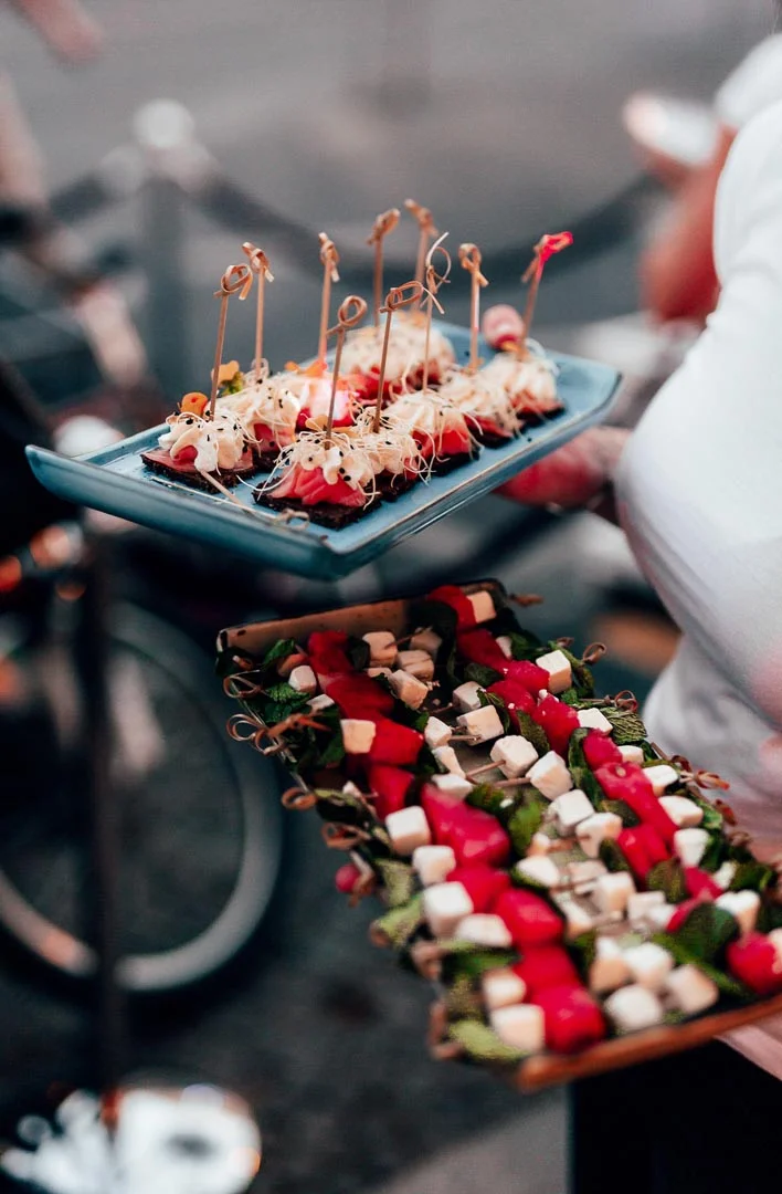 A person is holding two trays of assorted appetizers, perfect for any function room hire. The top tray features skewers with sprouts and red toppings, while the bottom tray showcases skewers with alternating pieces of cheese, watermelon, and greens. The background is blurred, featuring a bicycle. © Powerleague