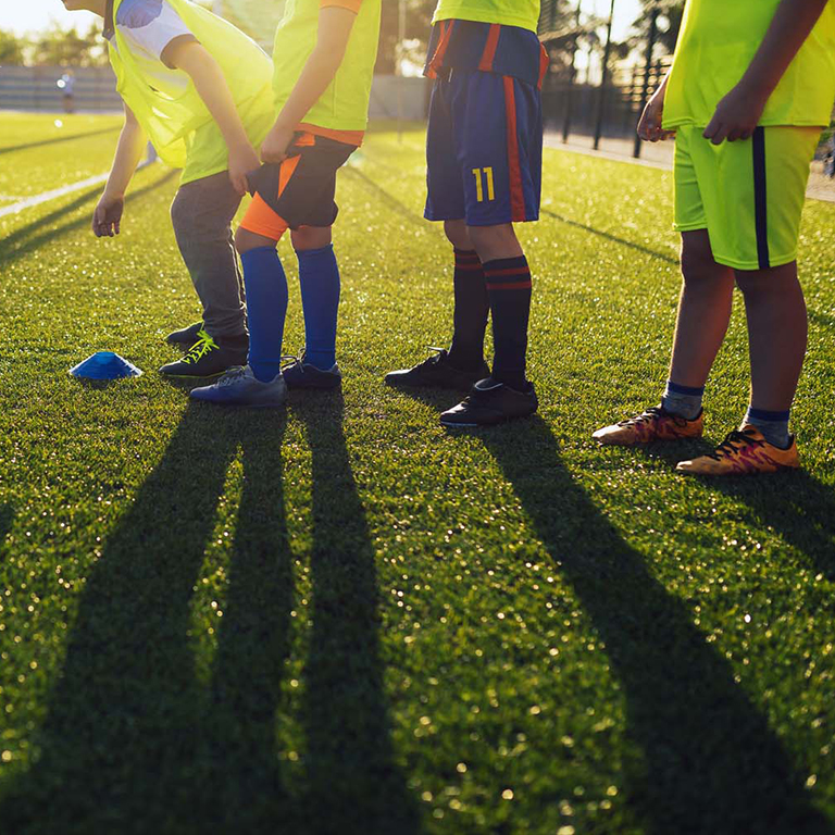 Children wearing sports attire and colorful bibs are lined up in a row on a sunlit artificial turf soccer field. Their shadows stretch long across the grass, and a blue cone marker is visible near their feet. Trees and a fence are in the background, capturing the essence of football holiday camps. © Powerleague
