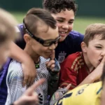 A group of children are huddled together, smiling and laughing in excitement. One child in the center wearing glasses stands out. They are all wearing colorful sports jerseys, suggesting a lively football birthday party in progress. The background is blurred, highlighting their joyful moment. © Powerleague