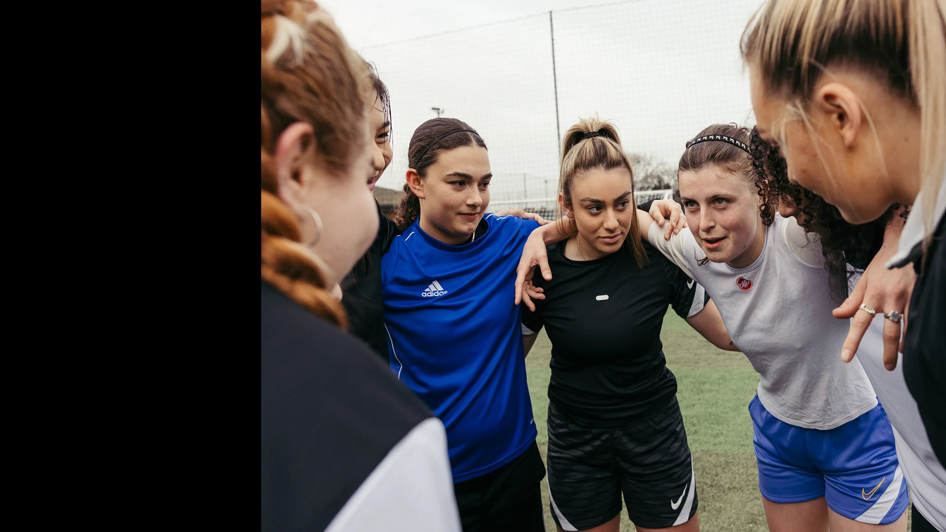 A group of young women in athletic wear huddled together on a sports field, preparing for a women's football match. They are putting their arms around each other's shoulders and appear to be listening and motivating each other before the game. The mood is focused and encouraging. © Powerleague