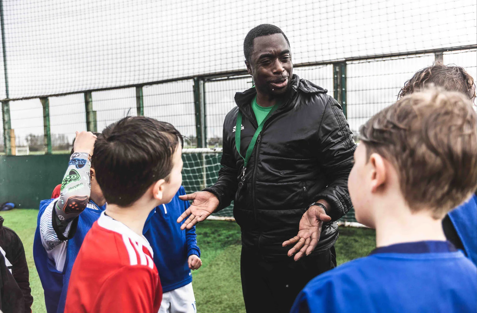 A coach wearing a black jacket with a green shirt underneath talks to a group of young soccer players during junior football training. The players, dressed in red and blue jerseys, stand around the coach on an outdoor field, listening attentively. In the background, a goal and fence are visible. © Powerleague