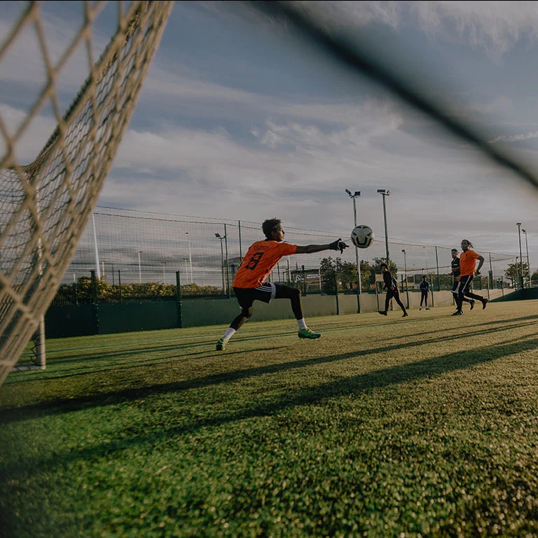 A soccer player in an orange jersey, possibly eyed by scouts in the transfer market, attempts to score a goal by kicking a ball toward the net. The goalkeeper reaches out with both arms to stop the ball. Other players can be seen in the background on the grassy field under a partly cloudy sky. © Powerleague