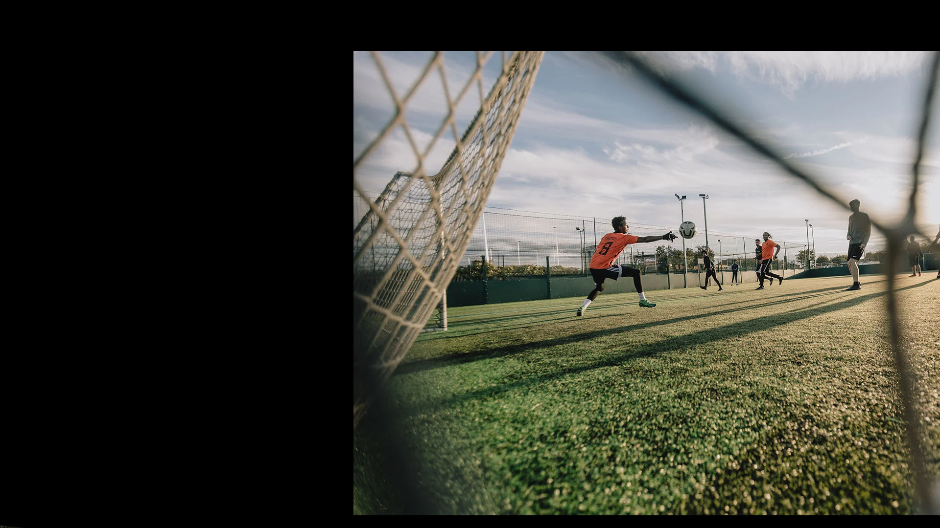 A soccer player in a red jersey, potentially eying the transfer market, attempts to block a goal, diving toward a ball mid-air, while other players are in action on the field under a partly cloudy sky. The image is framed by the net of the goalpost, with sunlight casting shadows on the grass. © Powerleague