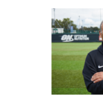 A man stands on a green soccer field with arms crossed, wearing a black England football coaching jacket and a watch, with a fence and trees in the background. The logo 