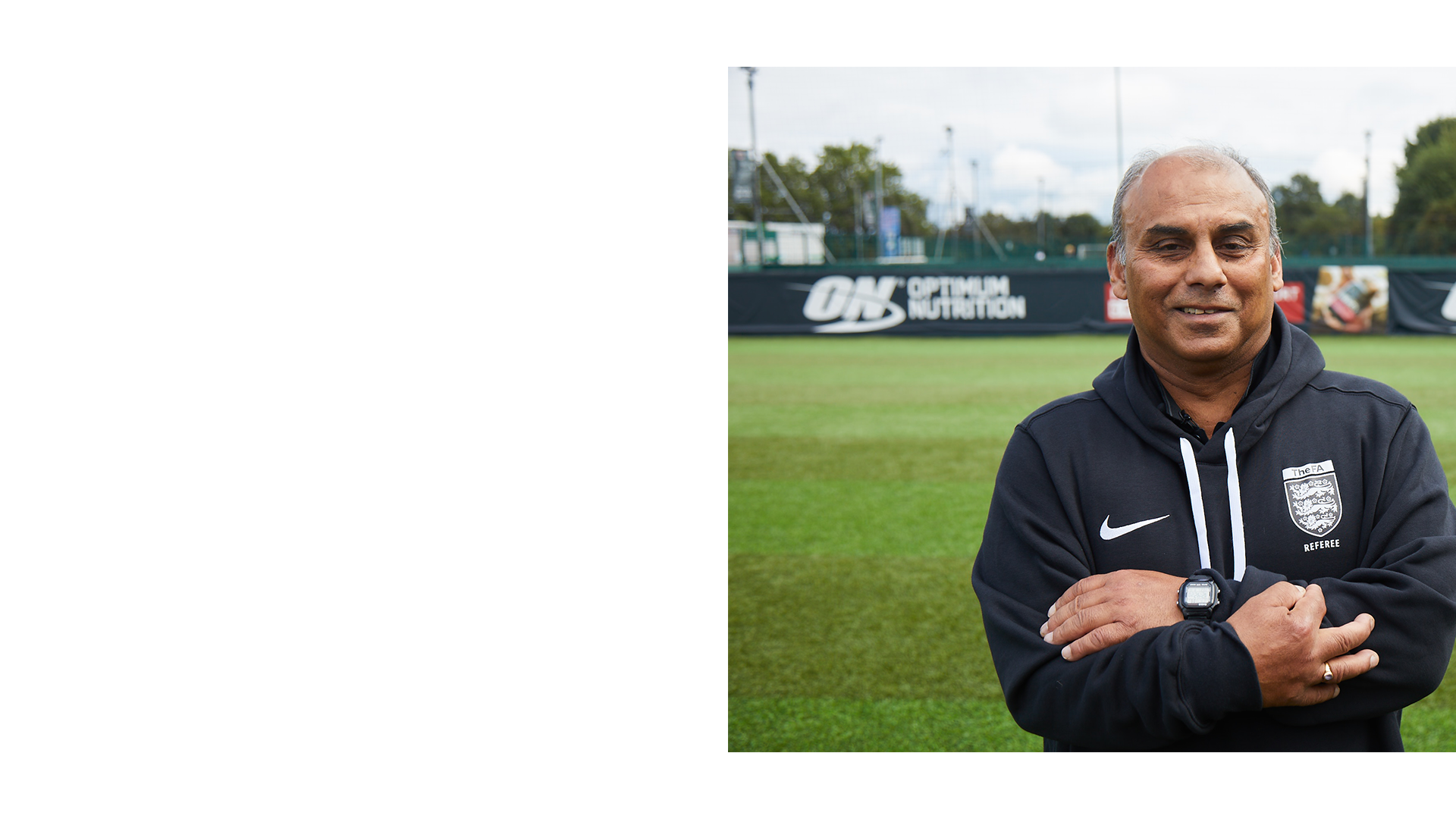 A man stands on a green soccer field with arms crossed, wearing a black England football coaching jacket and a watch, with a fence and trees in the background. The logo 