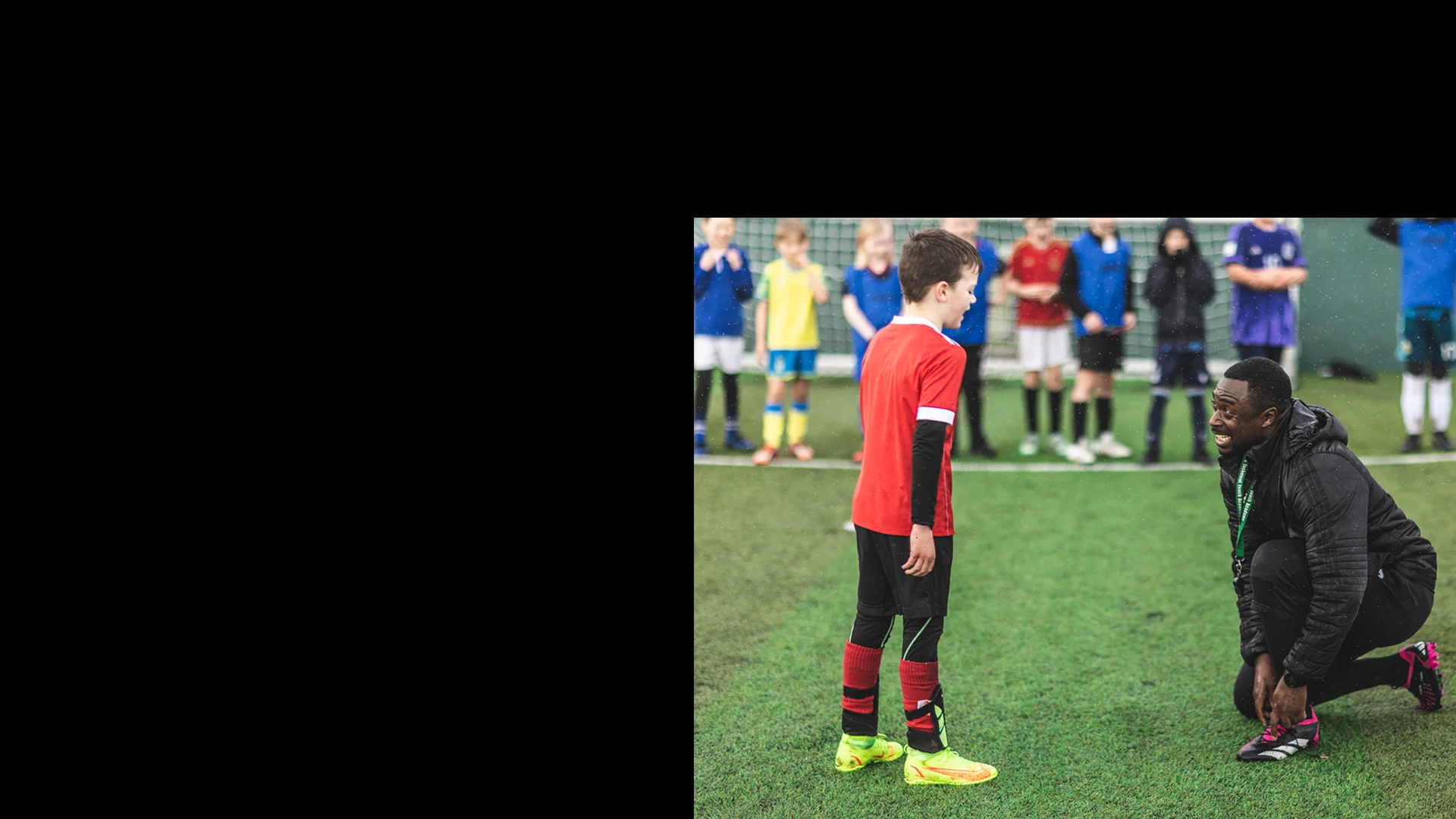 A soccer coach kneels on one knee, speaking to a young boy in a red jersey on a grassy field. In the background, other children in various colored jerseys stand and watch the interaction, indicating that this scene is part of a junior football training session. © Powerleague