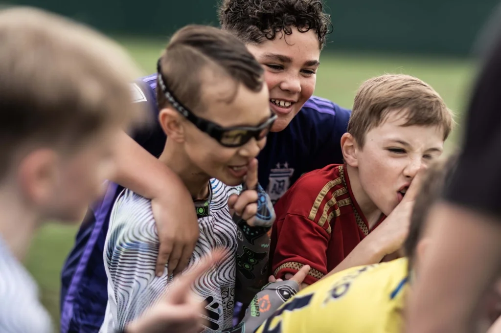A group of children, wearing sports jerseys, gather closely in a joyful huddle outdoors at one of the football holiday camps. One boy in the foreground is wearing sports glasses and smiling, while others are laughing and looking towards something out of frame. The group appears to be engaged and having fun. © Powerleague