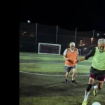 Three older adults enjoying a game of walking football on a well-lit artificial turf field at night. Two players in orange pinnies chase a player in a green pinny who is dribbling the ball towards the goal. A netted fence surrounds the field. © Powerleague