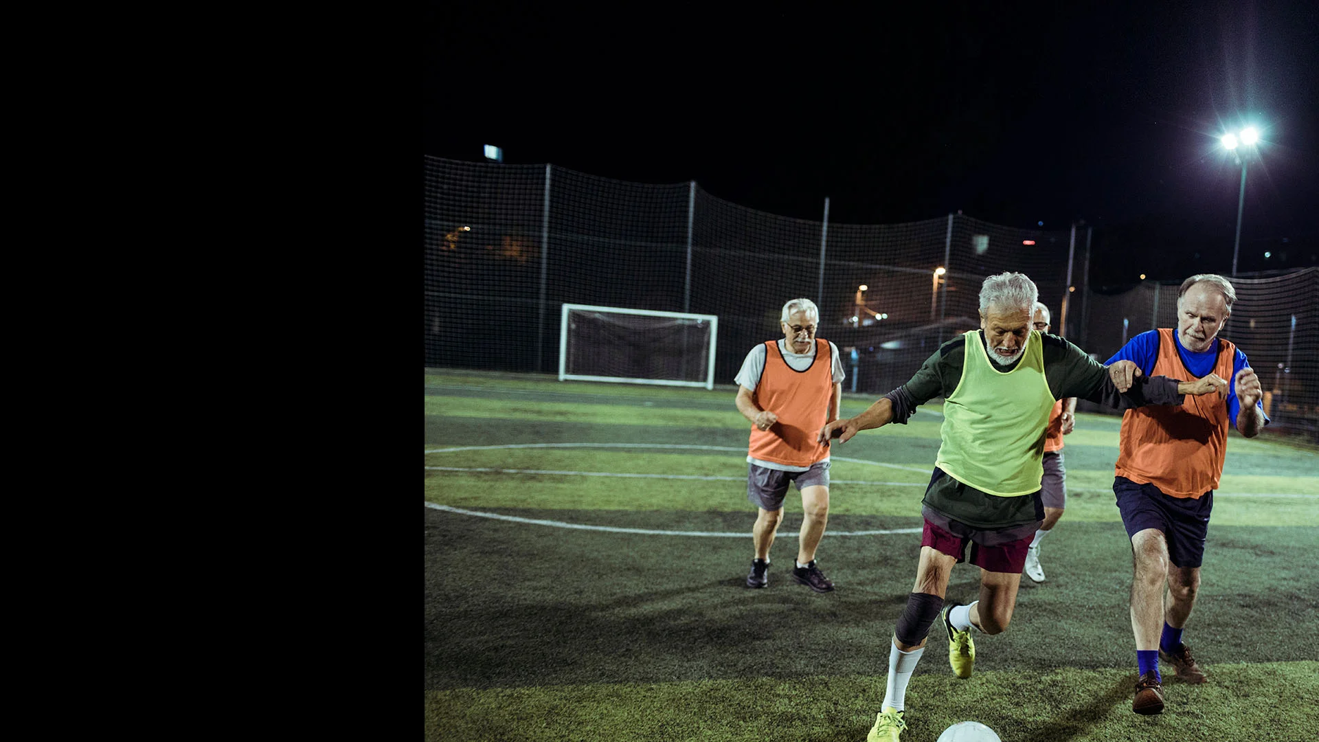 Three older adults enjoying a game of walking football on a well-lit artificial turf field at night. Two players in orange pinnies chase a player in a green pinny who is dribbling the ball towards the goal. A netted fence surrounds the field. © Powerleague