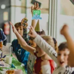 A group of children raise their arms in excitement at a football birthday party. They are sitting around a table with food, and one child is wearing a party hat. A sign in the background reads, 