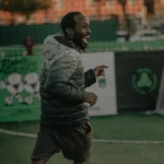 A man wearing a puffer jacket and shorts is joyfully running on a sports field, embodying health and vitality. He has a big smile on his face and appears to be playing a game or celebrating. The background shows banners and a backdrop of trees and buildings. © Powerleague