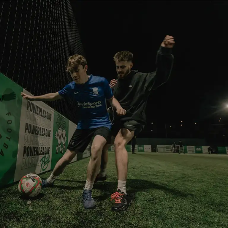 Two young men are playing soccer at night on an outdoor field. The player in the blue jersey is trying to shield the ball from an opponent in a black jersey. The field, surrounded by fences and lit by artificial lighting, is adorned with Powerleague banners showcasing advertising and partnerships. © Powerleague