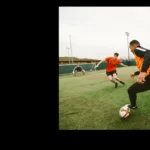 A soccer player in a black and orange jersey dribbles the ball, pursued closely by an opponent in a red jersey. Another player in red is running across the field towards the action. The game, reminiscent of intense football tournaments, is taking place on an outdoor turf field under a cloudy sky. © Powerleague