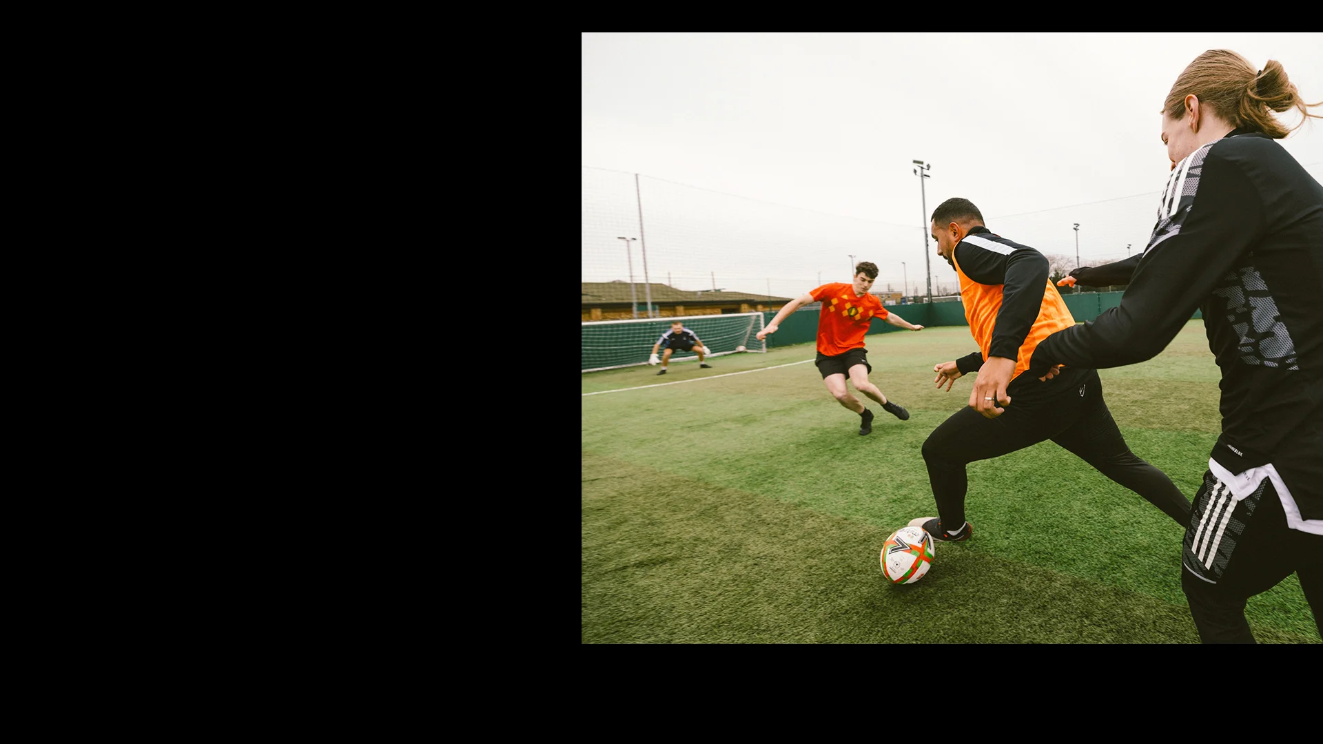 A soccer player in a black and orange jersey dribbles the ball, pursued closely by an opponent in a red jersey. Another player in red is running across the field towards the action. The game, reminiscent of intense football tournaments, is taking place on an outdoor turf field under a cloudy sky. © Powerleague