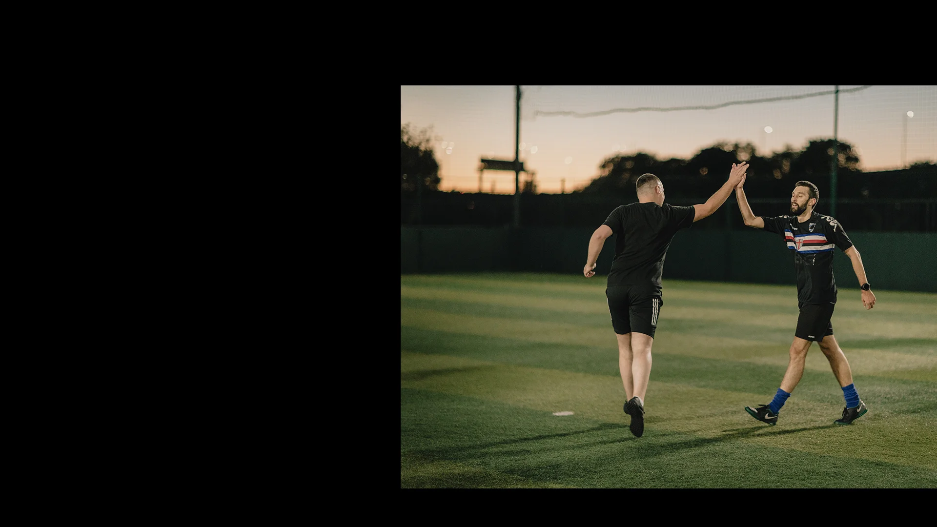 Two veterans in sports attire high-five on a soccer field at dusk. The man on the left is wearing black shorts and a black shirt, while the man on the right sports a striped jersey. The background reveals blurred trees against a warm sky transitioning to night. © Powerleague