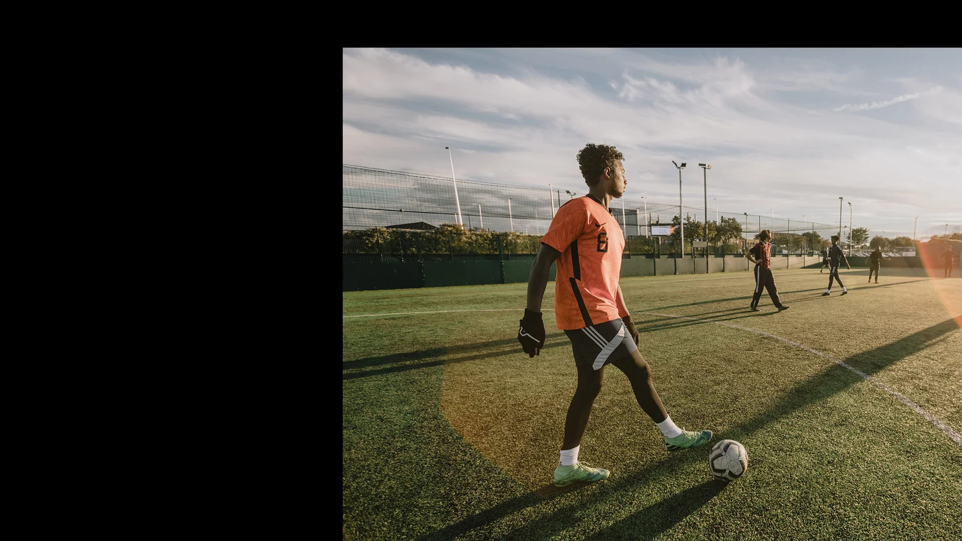 A soccer player in an orange jersey and green cleats touches the ball with one foot on an outdoor field, showcasing their athletic skill. They prepare to pass or kick the ball while other players and a fence are visible in the background. The sun is low, casting long shadows that emphasize the health benefits of outdoor activity. © Powerleague