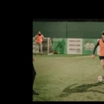 A soccer player in an orange training vest dribbles a ball on a green field during a night practice. Another player wearing a similar vest stands near the goal. The surroundings, complete with fencing and advertisements, highlight the vibrant atmosphere created by advertising and partnerships at Powerleague. © Powerleague