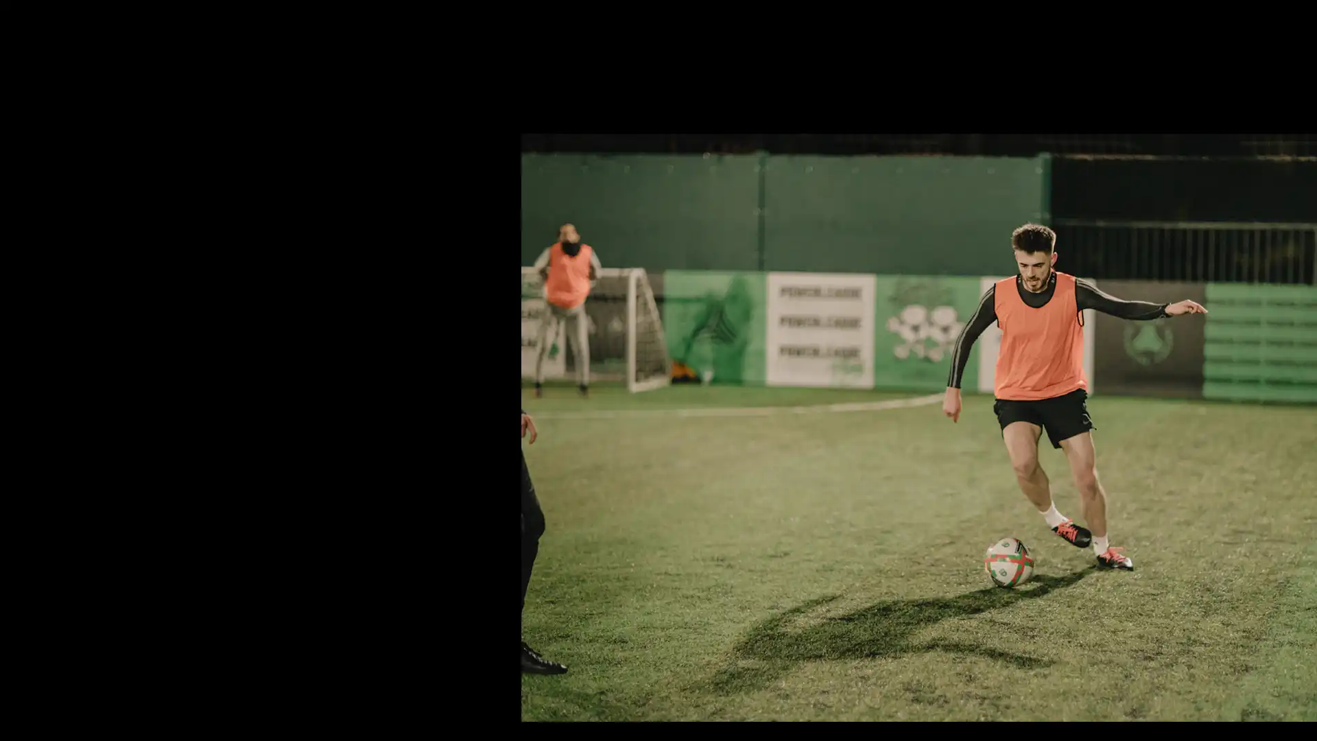 A soccer player in an orange training vest dribbles a ball on a green field during a night practice. Another player wearing a similar vest stands near the goal. The surroundings, complete with fencing and advertisements, highlight the vibrant atmosphere created by advertising and partnerships at Powerleague. © Powerleague