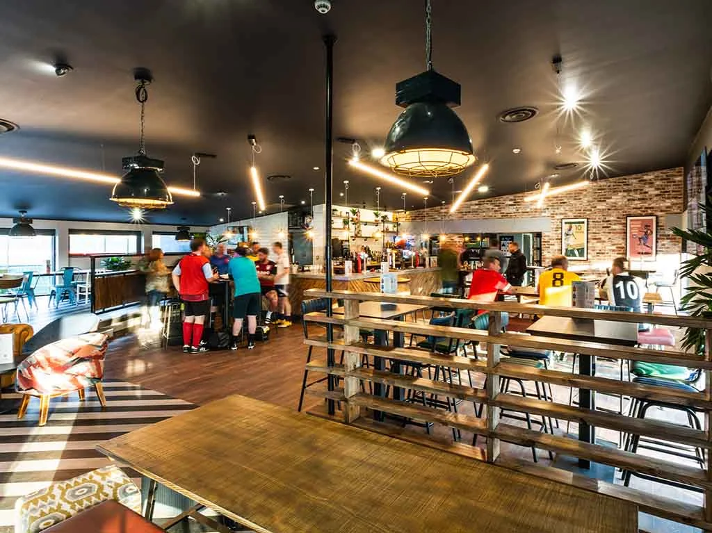 A modern, well-lit sports-themed bar with wooden furniture. Customers in sports jerseys gather near the bar, while others are seated at tables. The decor includes sport-related artwork and large overhead lights. The atmosphere appears lively and welcoming. © Powerleague