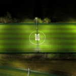 Aerial view of a well-lit, rectangular soccer field at night. The field has even green stripes running lengthwise with white lines marking the boundaries and center circle. Four tall floodlights illuminate the field. Surrounding areas are dark and grassy. © Powerleague