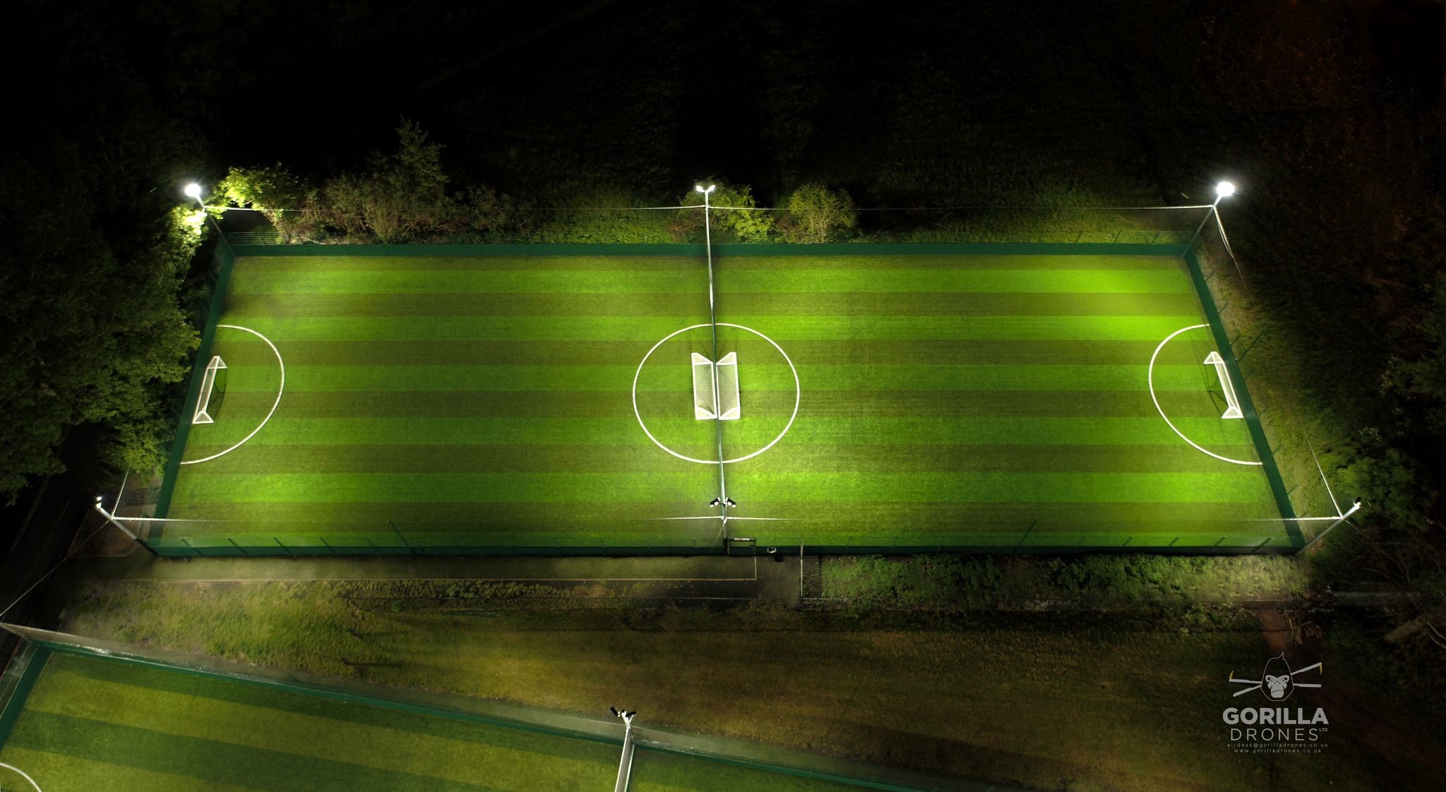 Aerial view of a well-lit, rectangular soccer field at night. The field has even green stripes running lengthwise with white lines marking the boundaries and center circle. Four tall floodlights illuminate the field. Surrounding areas are dark and grassy. © Powerleague
