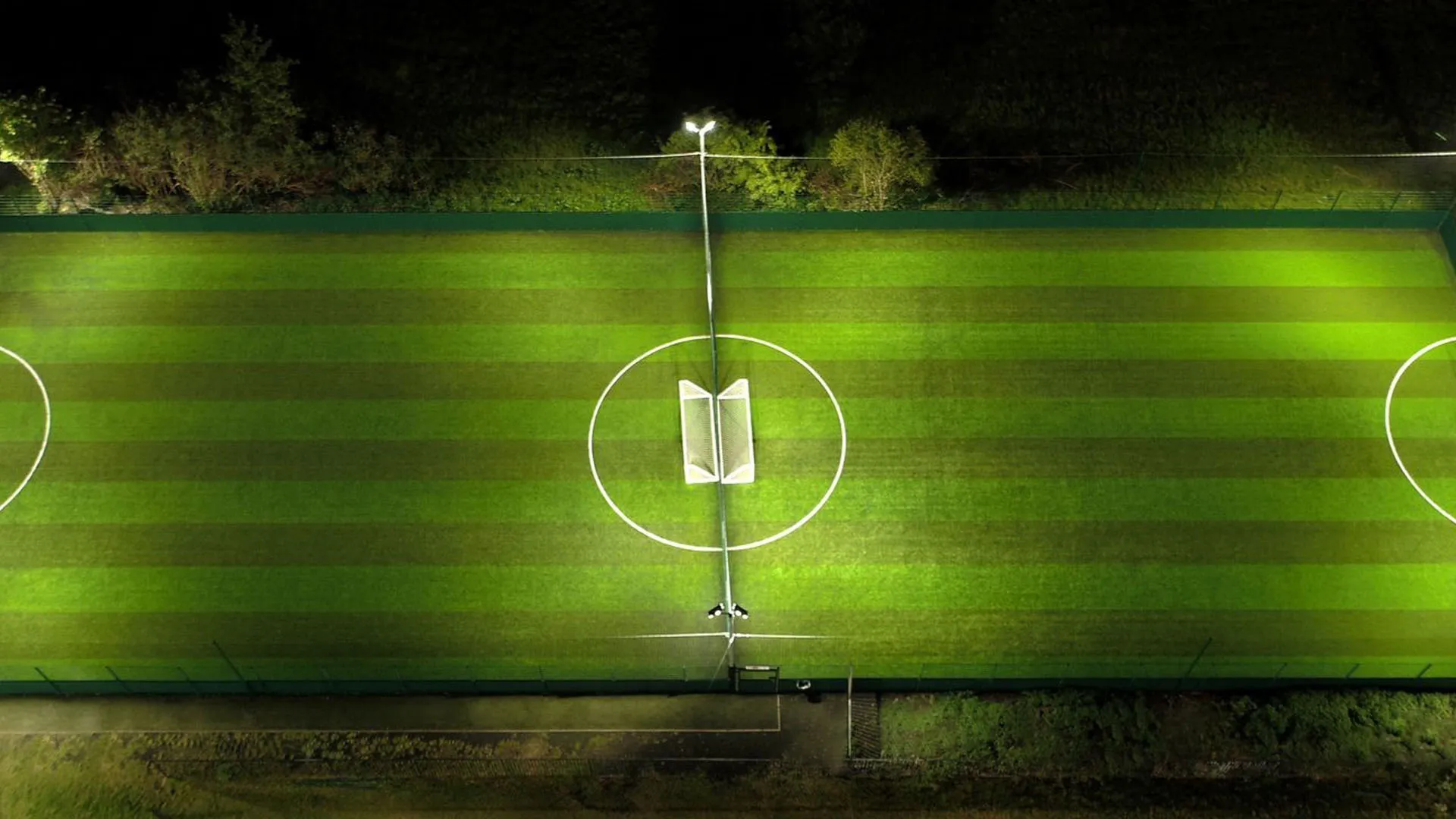 Aerial view of a well-lit soccer field at night. The neatly striped green grass is divided by white lines marking the playing area. The center circle contains a small structure, possibly equipment or a storage shed, illuminated by bright overhead lights. © Powerleague