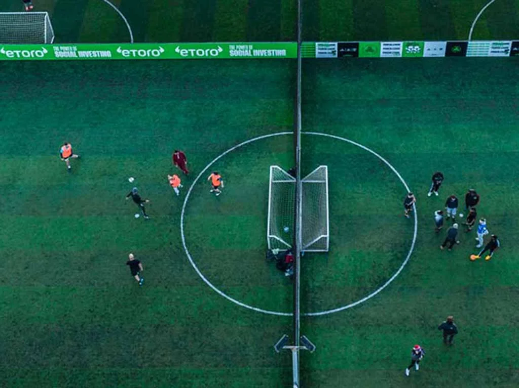 Aerial view of a soccer field with players in orange and dark jerseys. The field is divided by a net, and players are positioned on either side. Some players are near a goal positioned in the center of the field. Green advertisements line the top border of the field. © Powerleague