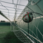 A soccer ball hitting the back of a green net, captured mid-flight, likely after scoring a goal. The ball has a white base with red and green accents, and the net is taut against the goalpost on a grass field, reminiscent of jubilant moments celebrated in any function room hire event. © Powerleague