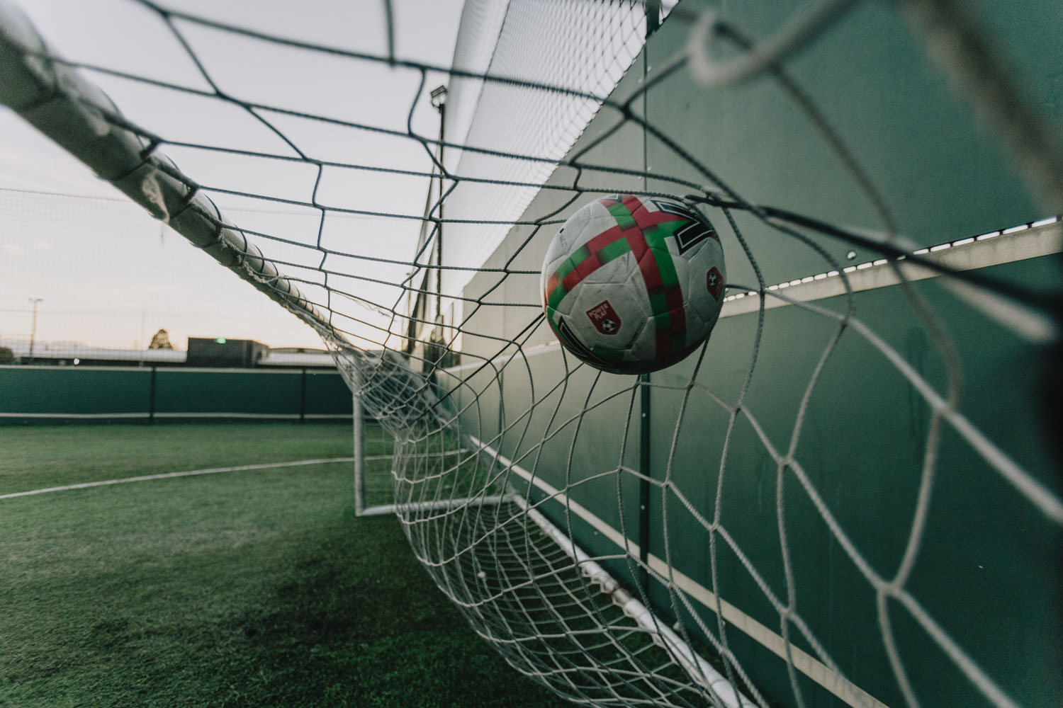 A soccer ball hitting the back of a green net, captured mid-flight, likely after scoring a goal. The ball has a white base with red and green accents, and the net is taut against the goalpost on a grass field, reminiscent of jubilant moments celebrated in any function room hire event. © Powerleague