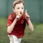 A young boy in a red and gold soccer jersey is celebrating a goal with enthusiasm during one of the football holiday camps. He is holding up both his hands with index fingers pointing upwards against a blurry, green field. © Powerleague