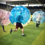 A group of people wearing inflatable bubble suits play a game of bubble football on an indoor field. One person, in a blue bubble suit, is running forward, while two others in clear suits follow. The field has green artificial turf and is surrounded by industrial-style walls and lighting. © Powerleague