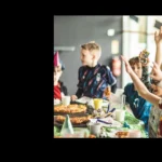 A group of children wearing party hats are gathered around a table filled with food, excitedly raising their hands. The scene is festive, resembling a football birthday party with a variety of snacks and drinks on the table. In the background, a sign reads 