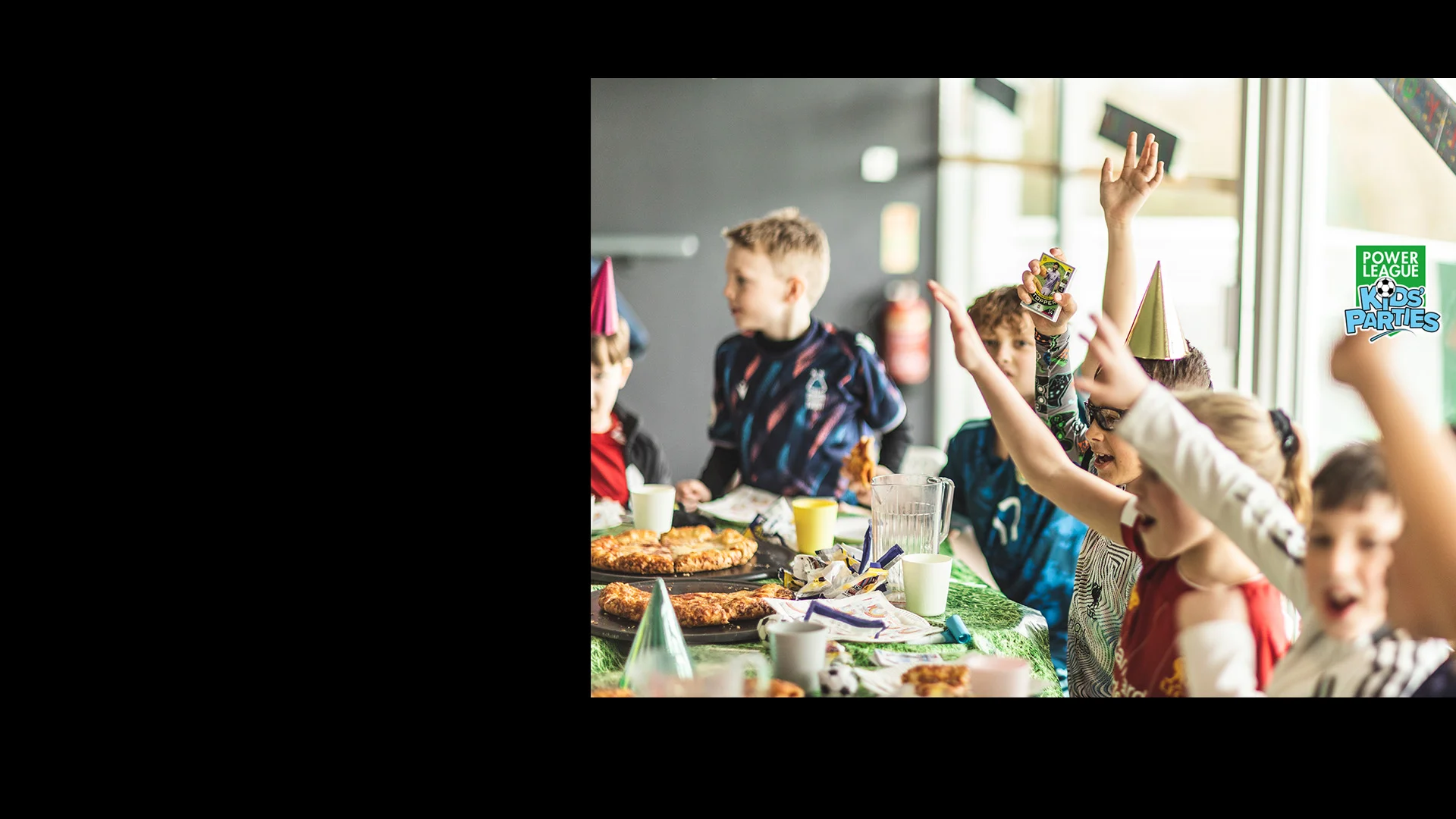 A group of children wearing party hats are gathered around a table filled with food, excitedly raising their hands. The scene is festive, resembling a football birthday party with a variety of snacks and drinks on the table. In the background, a sign reads 