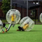 Four people are playing bubble football on a green field. They are inside large inflatable bubbles, with some players falling while others are upright. A brick building and trees are visible in the background. Everyone is wearing sports attire. © Powerleague