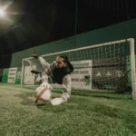 A soccer goalie in a gray outfit dives to the left, fully extending in an attempt to save a ball in front of a net. The scene is illuminated by bright lights, and there is green netting and advertising banners in the background. It appears to be nighttime, perfect for those looking to join a small-sided football league at Powerleague. © Powerleague