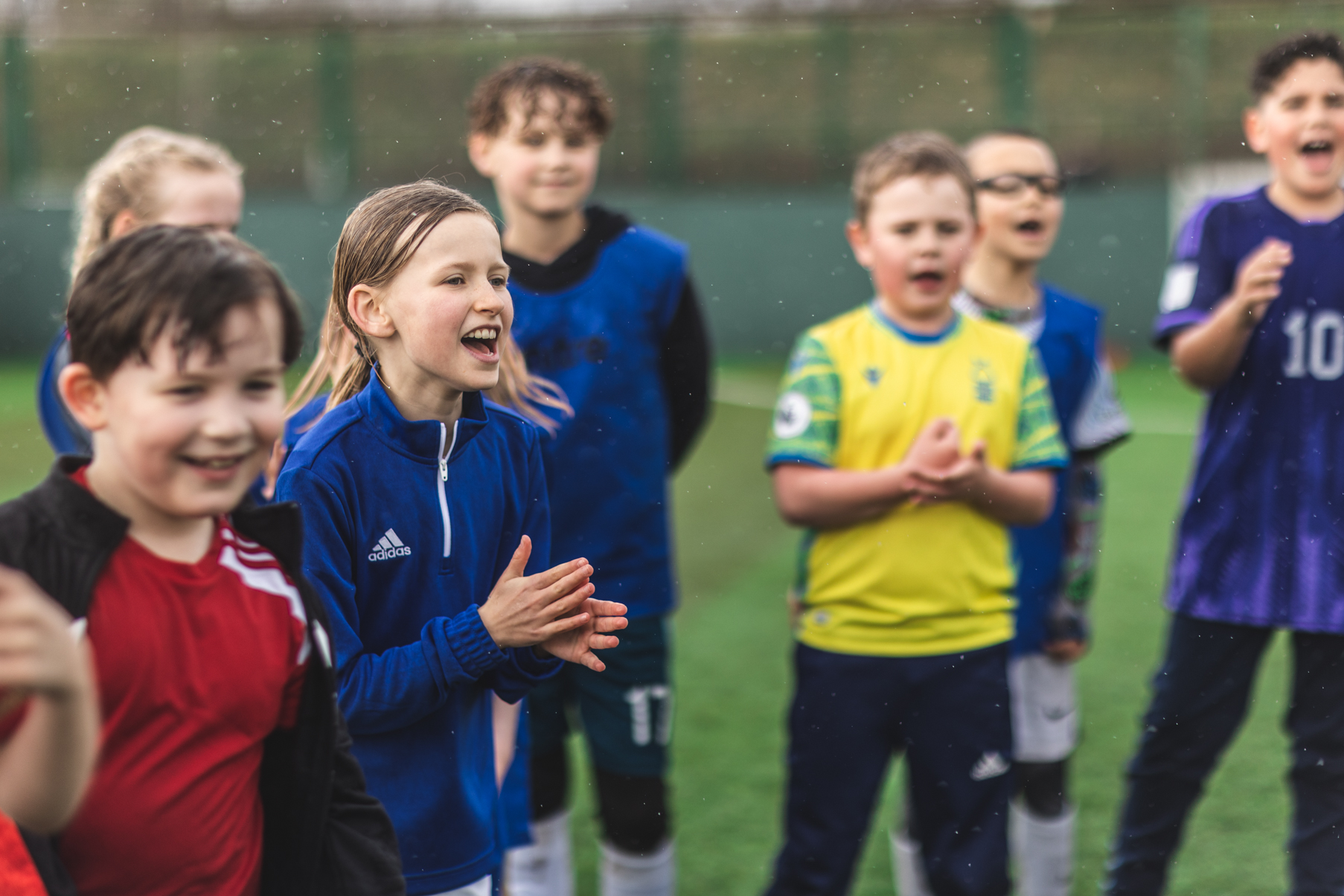 A group of young children stands on a soccer field, smiling and clapping. They are wearing various soccer jerseys and appear to be celebrating or cheering at the Powerleague Bradford opening event. The background shows a green fence and a slightly blurred landscape, indicating an outdoor setting. © Powerleague