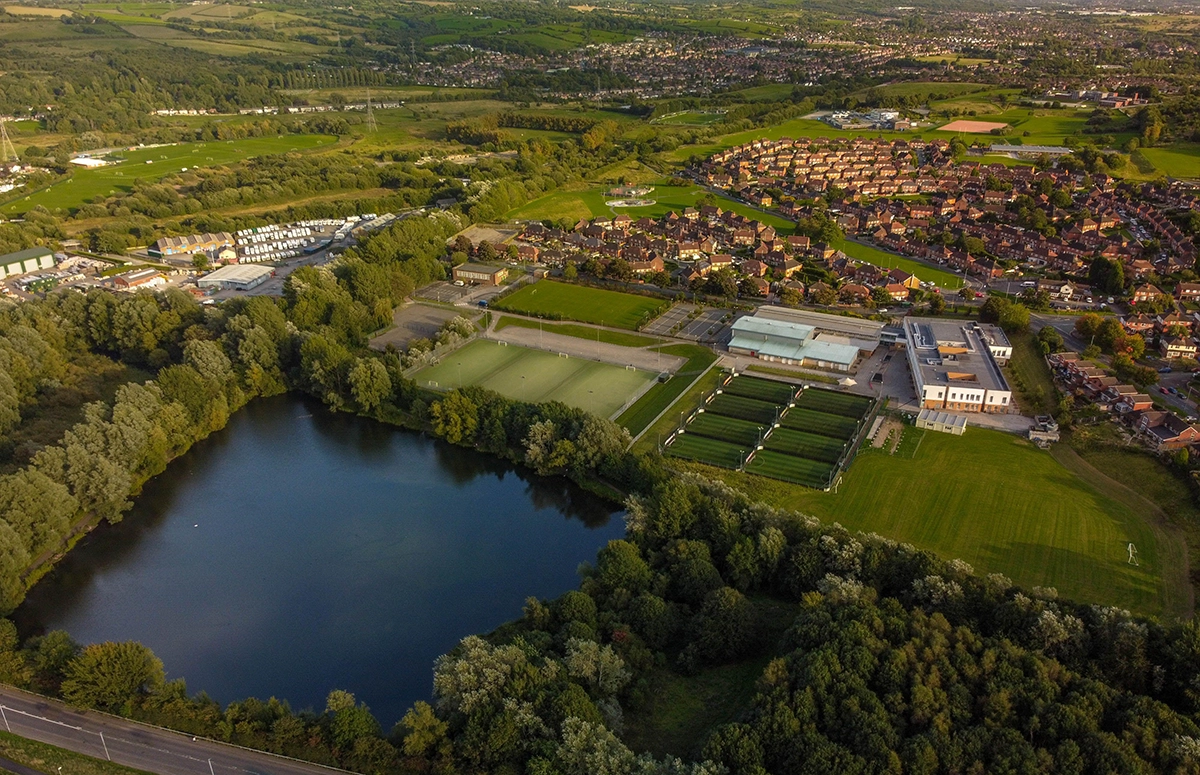 Aerial view of a calm lake surrounded by dense green trees and grass fields. Adjacent to the lake, there's a sports complex where you can join a small-sided football league at Powerleague, with several soccer fields and buildings. Nearby, a residential area with numerous houses and neatly organized streets is visible. Background shows vast green landscape and towns. © Powerleague