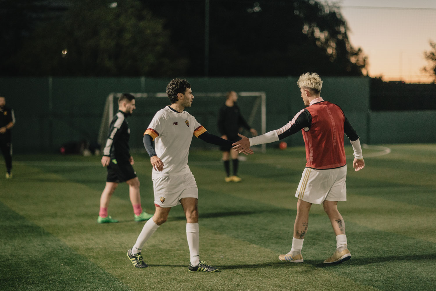 Two soccer players in white uniforms give each other a high-five on an outdoor field during dusk. In the background, other players and goalposts are visible. One player wears a red training vest, and the scene is lit by twilight. © Powerleague