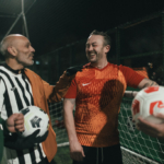 Four people are gathered on a soccer field at night, smiling and conversing. Two are wearing referee jerseys and holding soccer balls, while one in an orange jersey and another in a red jersey are chatting animatedly. The background features a fence and the faint lights from the Stoke Excel stadium. © Powerleague
