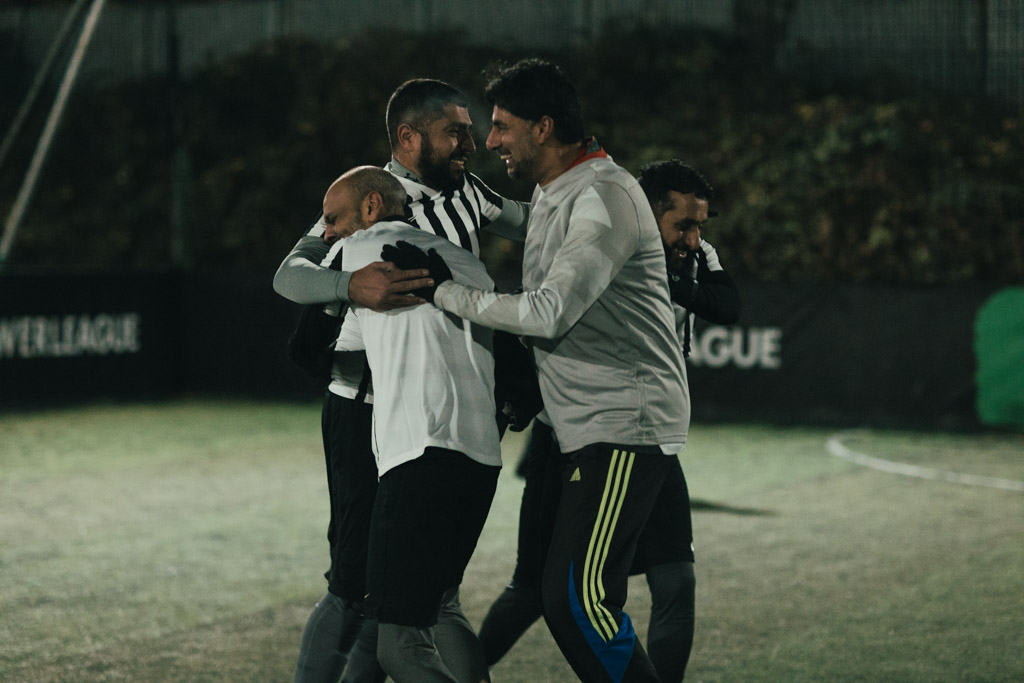 A group of four men wearing athletic clothing, including jerseys and shorts, are seen celebrating and hugging on an outdoor soccer field at night. The mood is festive, indicating they managed to stoke their camaraderie to excel in this moment of triumph. © Powerleague