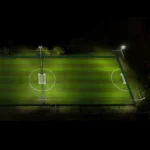 A nighttime overhead view of a well-lit soccer field. The field is empty, with perfectly manicured green grass and evenly spaced white goalposts at each end. The field is illuminated by four floodlights at the corners. © Powerleague