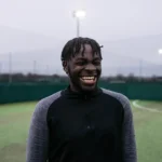 A person with dark skin and medium-length dreadlocks is standing on a soccer field at dusk, wearing a black and gray athletic top, and is smiling widely. The field, part of the 5-a-side voetbal voor iedereen at Powerleague Nederland, is surrounded by green netting with bright lights. © Powerleague