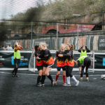 A group of women playing football. Four players in black jerseys with flame designs celebrate near the goalpost, while players in green vests prepare for the next play. The field is surrounded by netting, and there are colorful graffiti walls in the background. © Powerleague