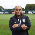 A man wearing a black jacket with a referee badge and a sports watch stands on a grass field with his arms crossed, embodying the authority of 5-a-side referees. The background shows a fence, advertisements, and some trees. © Powerleague