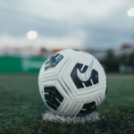 Close-up of a Nike soccer ball resting on a patch of artificial turf. The background is blurred, showcasing a field with goalposts and floodlights, suggesting an evening or early morning setting before a bubble football match or practice session. © Powerleague