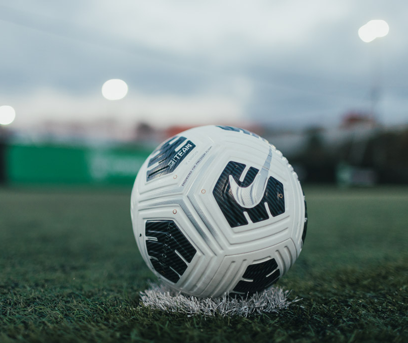 Close-up of a Nike soccer ball resting on a patch of artificial turf. The background is blurred, showcasing a field with goalposts and floodlights, suggesting an evening or early morning setting before a bubble football match or practice session. © Powerleague